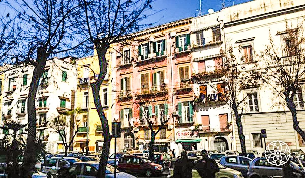 Rua arborizada de uma cidade italiana, com edifícios históricos de fachadas coloridas, lojas no térreo e carros estacionados ao longo da via.