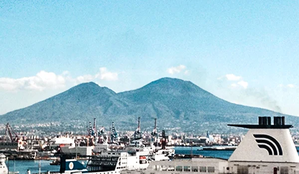 Vista do porto de Nápoles, com navios e estruturas portuárias em primeiro plano e o Monte Vesúvio ao fundo.