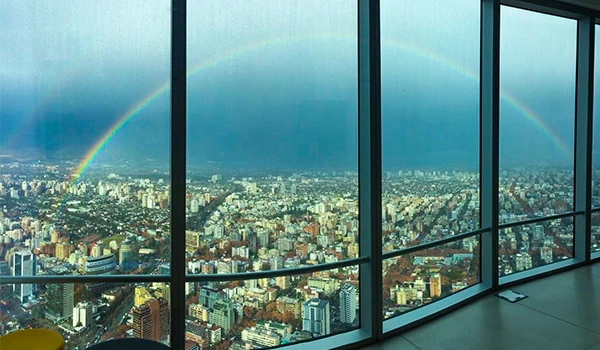 Vista de uma cidade maior de um prédio alto com janelas de vidro, arco-íris no céu e céu nublado ao fundo, criando uma paisagem urbana impressionante, do Sky Costanera.