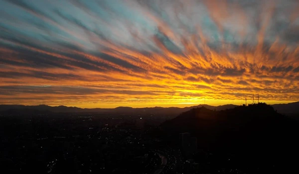 Belíssima foto de pôr do sol com nuvens coloridas no céu, destacando tons de laranja, rosa e azul, sobre uma paisagem montanhosa ao entardecer.