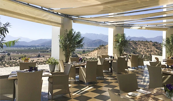 Varanda de restaurante com vista para as montanhas, mesas e cadeiras elegantes, ambiente ao ar livre com cobertura retrátil, plantas decorativas e ambientação moderna, ideal para refeições ao ar livre. Vinícolas no Chile, qual visitar?