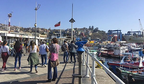 Imagem de um porto com pessoas caminhando na orla, barcos atracados e vista para o mar ao fundo, sob céu claro e ensolarado.
