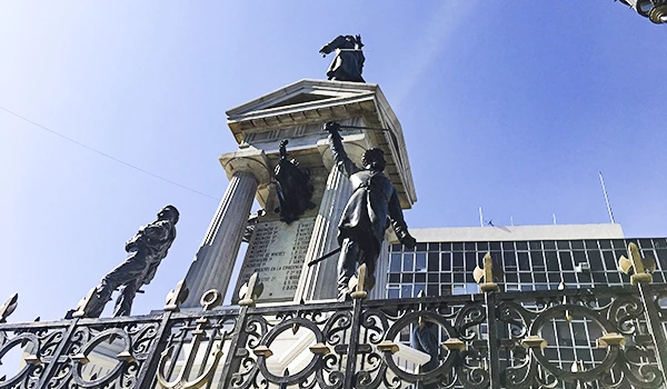 Imagem de esculturas em uma praça ao ar livre, com uma estrutura arquitetônica clássica ao fundo, sob um céu claro. As esculturas representam figuras humanas em diferentes posições.