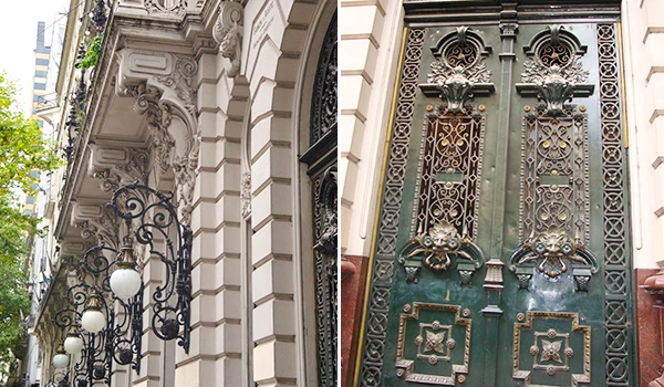 Imagem de uma porta de ferro com detalhes tradicionais e uma fachada de prédio antigo com ornamentos arquitetônicos elaborados em estilo clássico, em uma rua urbana.