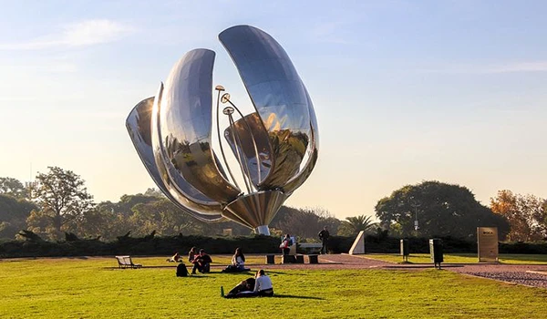 Escultura moderna de uma flor metálica gigante em um parque, com pessoas sentadas e caminhando ao redor, sob céu ensolarado.
