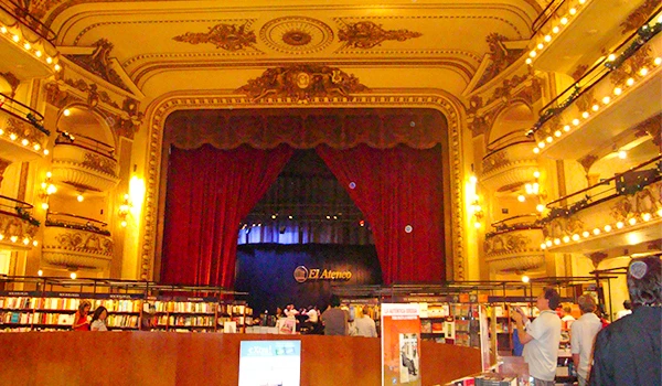 Foto do interior de uma biblioteca ou espaço cultural com palco e estantes cheias de livros, iluminação quente e pessoas explorando o local. Viagem para Buenos Aires.