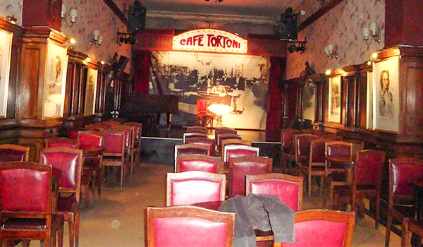 Interior de uma sala de show ou bar com cadeiras vermelhas, palco com piano, decoração vintage e iluminação suave, perfeito para noites de entretenimento.