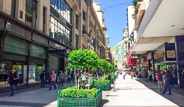 Rua movimentada com lojas comerciais, árvores decorativas e pessoas caminhando em uma cidade moderna e vibrante ao ar livre