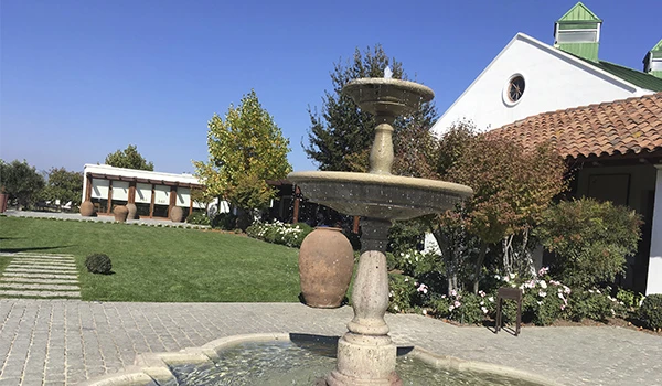 Imagem de uma fonte decorativa em um jardim bem cuidado, com árvores, grama bem aparada e uma construção ao fundo, ideal para ambientes ao ar livre.