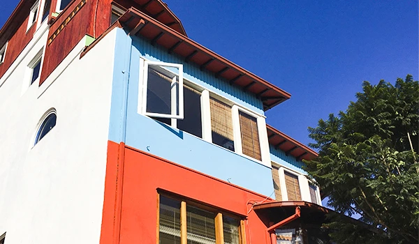 Imagem de fachada de uma casa com cores vibrantes, varanda e janelas grandes, cenário urbano com céu azul e árvores ao fundo.