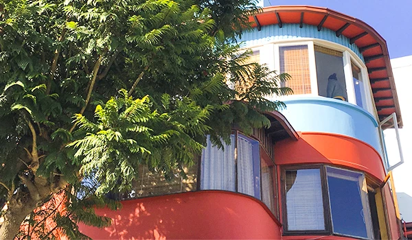 Imagem de Pablo Neruda com arquitetura colorida, destacando suas janelas e detalhes em vermelho e azul, sob céu claro, rodeado por árvores verdes.