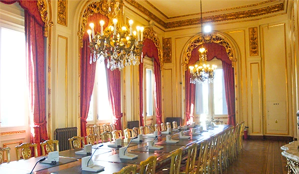 Salão de reuniões elegante e luxuoso com muita luz natural, cortinas vermelhas e móveis dourados, ideal para eventos corporativos e encontros formais.