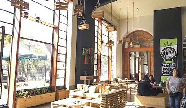 Interior de um restaurante com decoração rústica e mesas de madeira, luz natural entrando pelas grandes janelas, clientes sentados e ambiente acolhedor.
