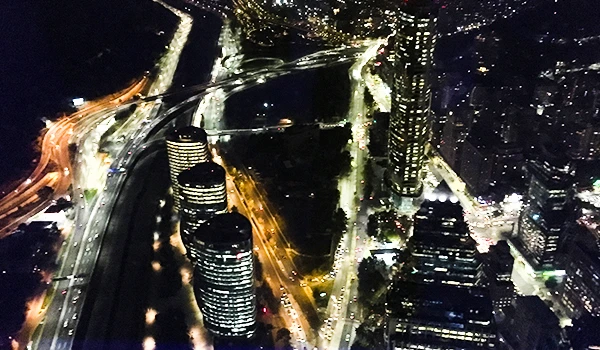 Imagem aérea de uma cidade moderna à noite, com edifícios altos iluminados, ruas movimentadas e um rio ao centro, destacando a vida urbana vibrante.