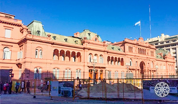 Imagem do Palácio do Governo de Buenos Aires, um edifício histórico rosa localizado na Argentina, com pessoas na frente e área de construção, sob um céu azul claro.