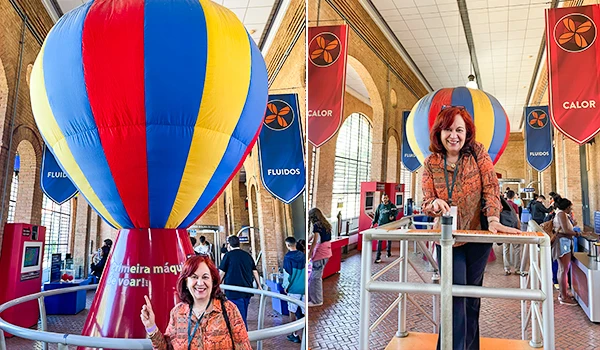 Deusa Rodrigues em ambiente indoor com balão de aquecimento de ar quente grande e colorido ao fundo, envolvendo a pessoa. Cenário com estruturas de suporte, banners promocionais e várias pessoas ao redor, em um evento ou feira.