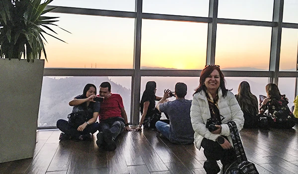 Grupo de pessoas sentado próximo a janela de um prédio durante o pôr do sol, algumas tirando fotos e conversando em um ambiente moderno e iluminado, no famoso Sky Costanera.