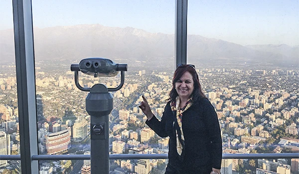 Deusa Rodrigues em observatório no topo de arranha-céu com vista panorâmica da cidade e montanhas ao fundo, usando jaqueta escura e acessório na roupa. Sky Costanera altura.