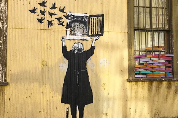 Street art de uma mulher com cabelo grisalho, vestindo um casaco preto, soltando pássaros negros de uma gaiola na parede amarela, simbolizando liberdade. Grafites de Valparaíso.