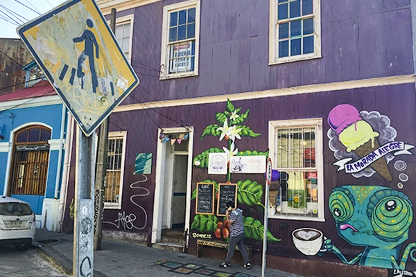 Rua com uma fachada colorida e grafitada, com um painel de arte de rua de uma criatura com olhos grandes e traços vibrantes, com uma placa de trânsito e pessoas andando. Grafites de Valparaíso.