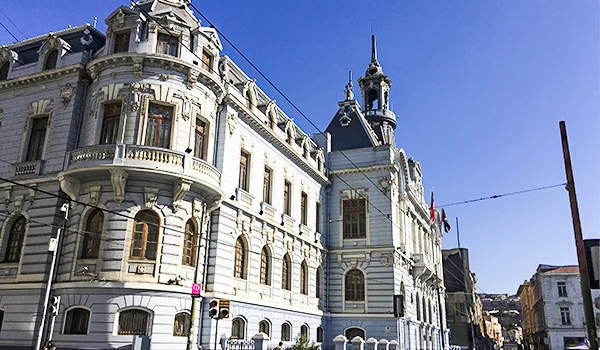 Edifício histórico de arquitetura clássica na cidade, com detalhes ornamentais, janelas grandes e torres, sob céu azul claro.