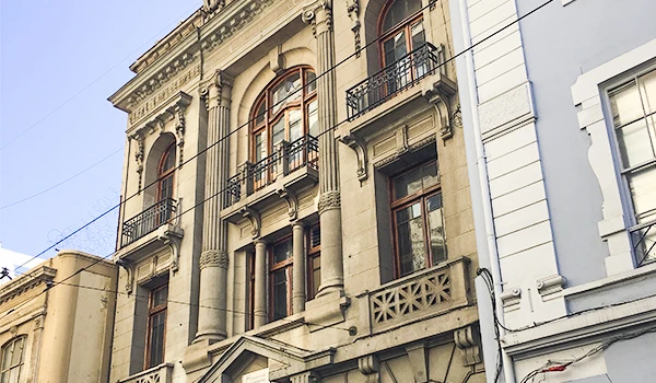 Imagem de uma fachada de edifício histórico com arquitetura clássica, detalhes ornamentais e janelas grandes, localizado em uma rua urbana.