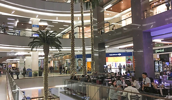 Imagem de um shopping center moderno com várias lojas, bancos e pessoas fazendo compras, decorado com árvores decorativas e iluminação aconchegante, destacando o ambiente comercial e de lazer.