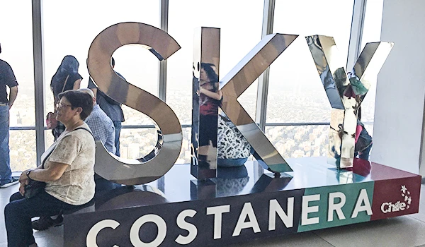 Imagem de turistas observando uma instalação com as letras 'SKY' Costanera, de uma altura grande, em uma atração turística com vista panorâmica da cidade ao fundo.