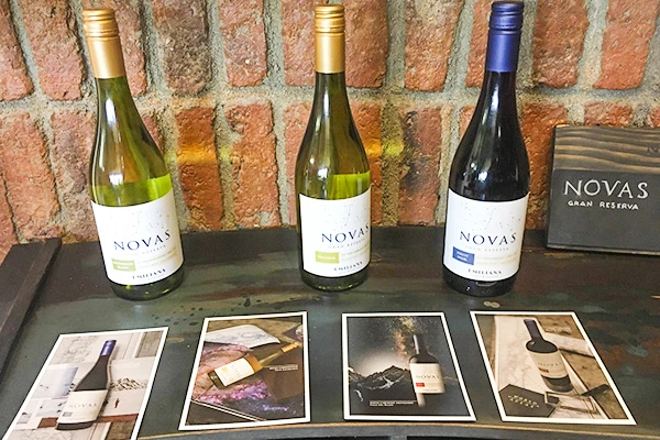 Três garrafas de vinho Novas Reserva variadas na cor, exibidas sobre uma mesa com fotos promocionais e uma parede de tijolos ao fundo. Vinícolas no Chile, qual visitar?