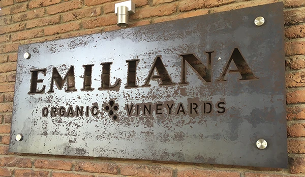 Placa de metal com o nome Emilliana Organic Vineyards em destaque na fachada de uma vinícola, com fundo de parede de tijolos expostos.