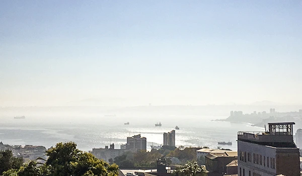 Vista panorâmica de uma cidade com mar ao fundo, céu ensolarado e alguns prédios baixos, refletindo um dia de clima claro e tranquilo.