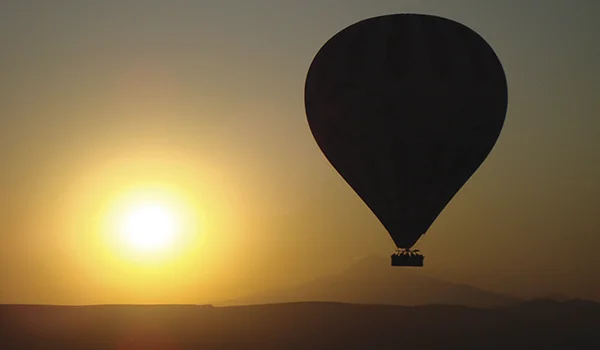 Balão de ar quente em silhueta ao pôr do sol, com o sol brilhando próximo ao horizonte e uma paisagem de montanhas ao fundo.