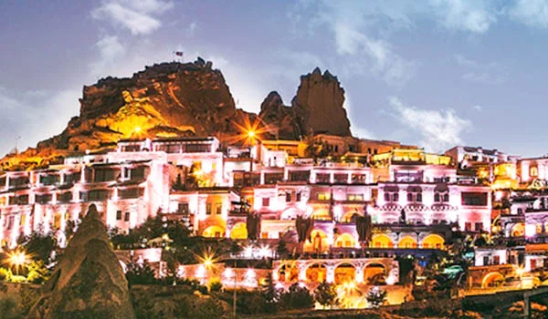 Paisagem noturna da Capadócia, na Turquia, com construções de arquitetura tradicional iluminadas por luzes amarelas e alaranjadas, erguidas ao pé de grandes formações rochosas. Ao fundo, destacam-se imponentes montanhas de pedra sob um céu azul-acinzentado, criando um cenário turístico pitoresco e encantador.