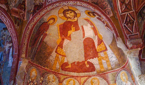 Afresco religioso em uma igreja rupestre da Capadócia, na Turquia, representando Jesus Cristo sentado em um trono, com auréola ao redor da cabeça, vestes em tons de vermelho e branco e figuras angelicais ao seu redor. A pintura apresenta cores terrosas e detalhes ornamentais, emoldurada por arcos e motivos decorativos medievais nas paredes e no teto de pedra.