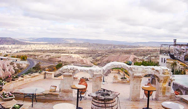 Texto alternativo: Terraço de hotel na Capadócia, na Turquia, com mesas ao ar livre, paredes de pedra e vista panorâmica das formações rochosas e vales da região sob um céu nublado.
