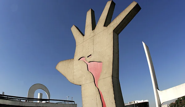 Monumento de concreto em forma de mão, com o mapa da América Latina pintado em vermelho na palma. Ao fundo, há um céu azul, uma estrutura metálica e parte de um complexo arquitetônico.
