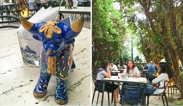 Fotografia dividida em duas cenas: à esquerda, uma escultura decorativa de um touro azul com detalhes dourados e coloridos sobre uma mesa de restaurante; ao fundo, há cadeiras, mesas e um cardápio. À direita, um grupo de pessoas está sentado em mesas ao ar livre, em um restaurante cercado por árvores e vegetação.