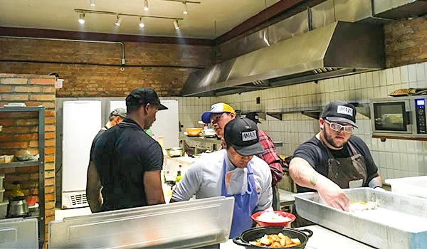  Cozinha de restaurante com vários cozinheiros preparando pratos. Ao fundo, há paredes de tijolos aparentes, azulejos brancos e uma grande coifa de metal. Na frente, dois cozinheiros usam aventais e bonés pretos, enquanto outros trabalham ao redor de bancadas e equipamentos de cozinha do Amazo restaurante peruano.