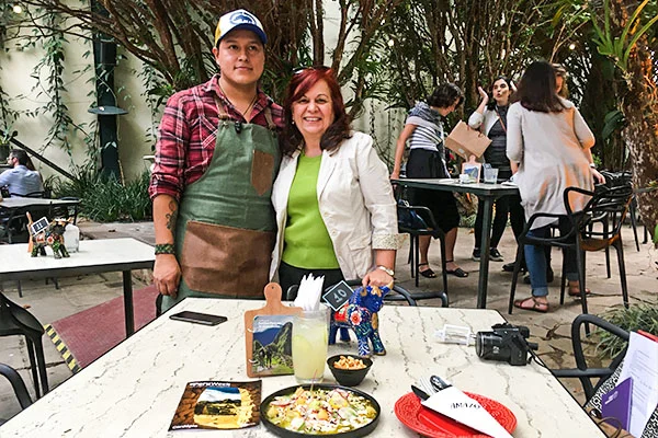 Deusa Rodrigues e o Chef posam diante de uma mesa com pratos de comida, bebidas e objetos decorativos em um restaurante ao ar livre. Uma delas usa avental verde e camisa xadrez; a outra veste uma jaqueta branca e blusa verde. Amazo restaurante peruano.