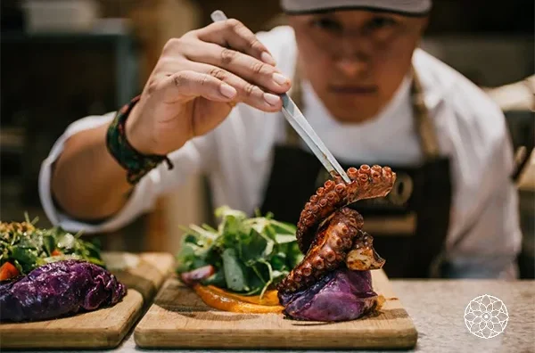 Chef preparando um prato de polvo grelhado com legumes e verduras sobre uma tábua de madeira, em um restaurante.