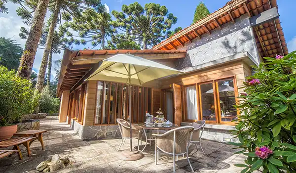 Imagem de uma casa de campo aconchegante com área externa, mesas e cadeiras ao ar livre, rodeada por árvores e plantas, ideal para relaxar e aproveitar a natureza.