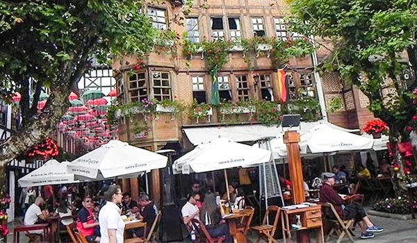Cenário de um restaurante na rua ao ar livre com muitas pessoas, mesas com guarda-sol branco e edifícios históricos ao fundo, rodeado de árvores verdes.