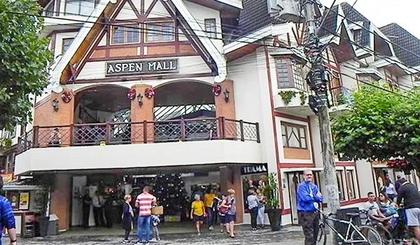 Fachada do Aspen Mall em uma rua movimentada, com várias pessoas entrando e saindo do shopping, cercada por árvores e postes de eletricidade.