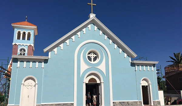 Igreja de cor azul claro com detalhes brancos e uma cruz no topo, situada em um ambiente ensolarado, com céu azul ao fundo, representando uma igreja católica.