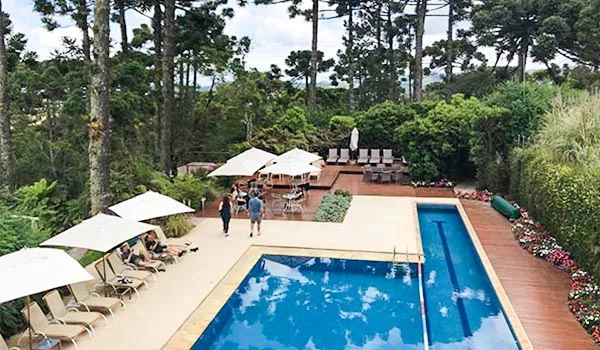 Piscina ao ar livre em espaço de lazer com espreguiçadeiras, guarda-sóis e área de descanso, rodeada por árvores e vegetação exuberante.