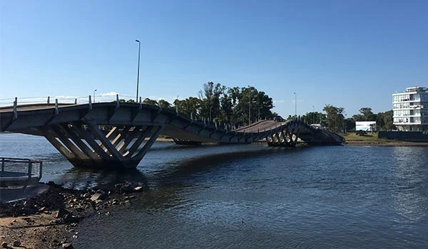 Imagem de uma ponte suspensa danificada sobre o rio, com partes do tabuleiro deslocadas e estrutura exposta, indicando possível dano ou acidente.