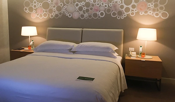 Quarto de hotel moderno com decoração elegante, cama de casal com roupas de cama brancas, abajures ao lado da cama e parede decorada com círculos artísticos.