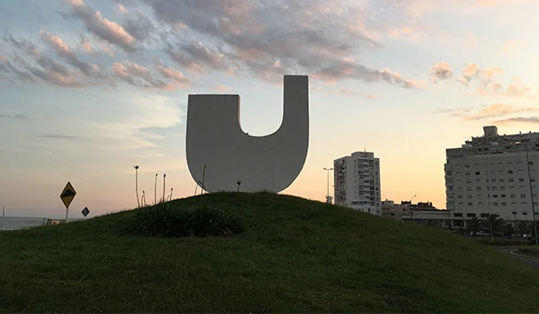 Escultura moderna em forma de letra U em uma praça, com prédios ao fundo ao entardecer, criando uma paisagem urbana e artística.