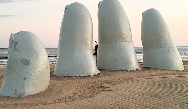 Escultura de conchas gigante na praia ao pôr do sol, com areia ao redor e céu com tonalidades suaves.