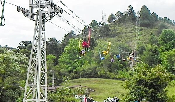 Diversas pessoas se aventuram em um passeio de cadeira de rodas na tirolesa em meio à natureza, com árvores verdes, morros e céu nublado ao fundo. Natal em Campos do Jordão.